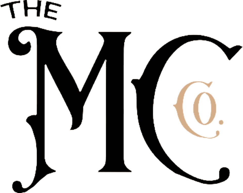 Stylized text "THE MC" in black on a beige background. "THE" is smaller and positioned above the left part of the letter "M".